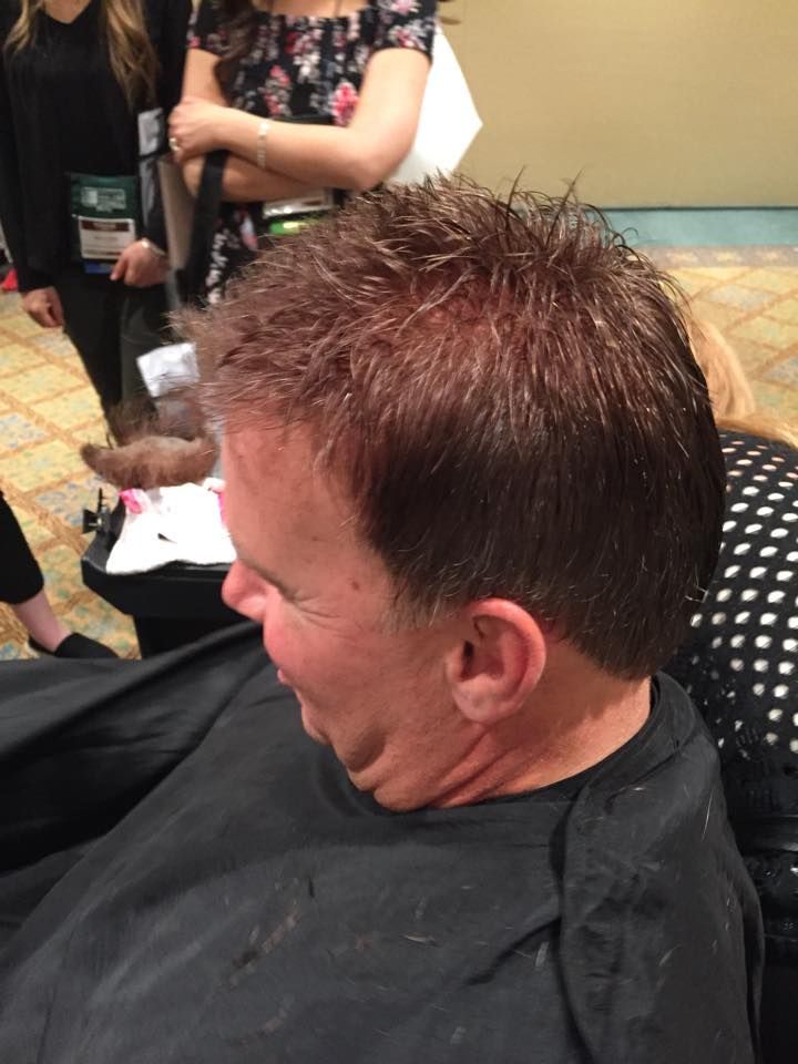Man getting a haircut; brown hair, smiling. Black cape. Indoors with people in background.