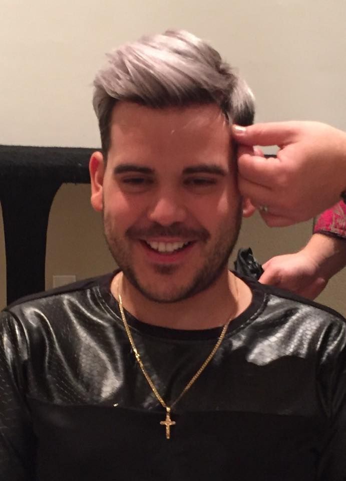 Man with silver hair, smiling, being touched by a hand. Wearing black shirt and gold cross necklace.