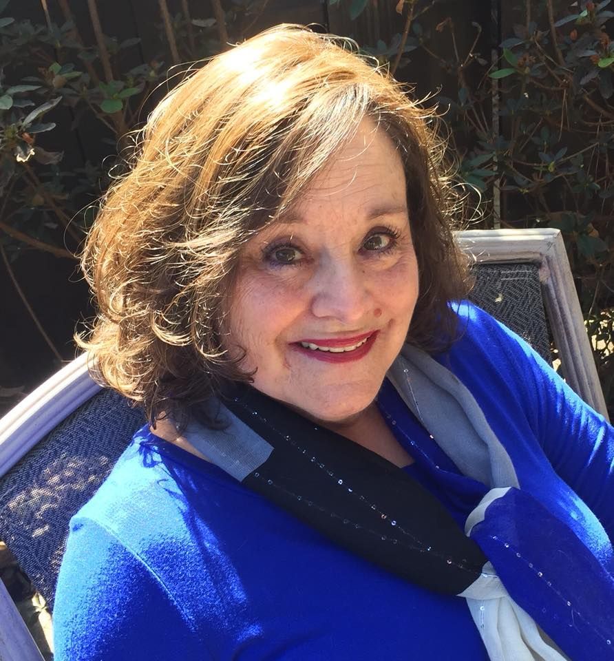 Woman with brown hair and red lipstick smiles, wearing a blue shirt and scarf, outside.