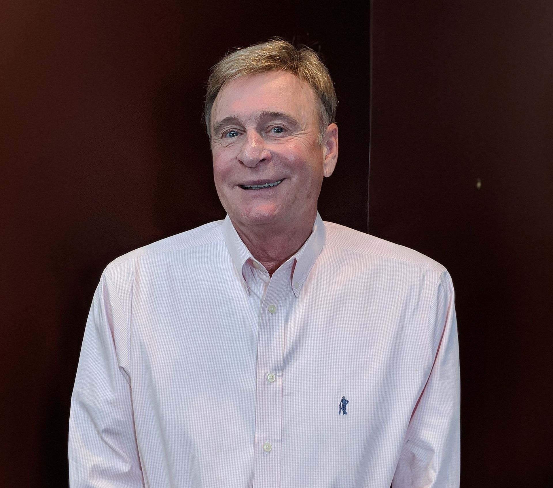 Smiling older man in a pink and white striped shirt, in front of a dark wall.