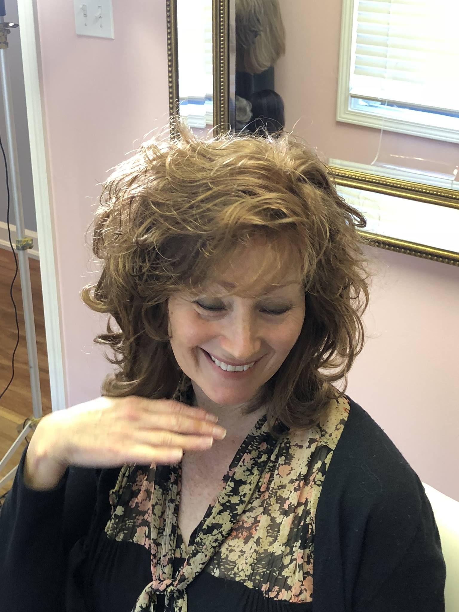 Woman with curly brown hair smiling, hand near face, wearing a black top with a scarf.