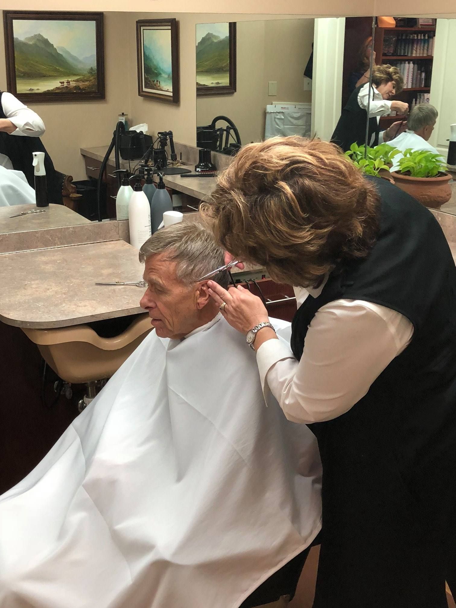 Barber cutting a man's hair in a salon. The man is seated and wearing a white cape.