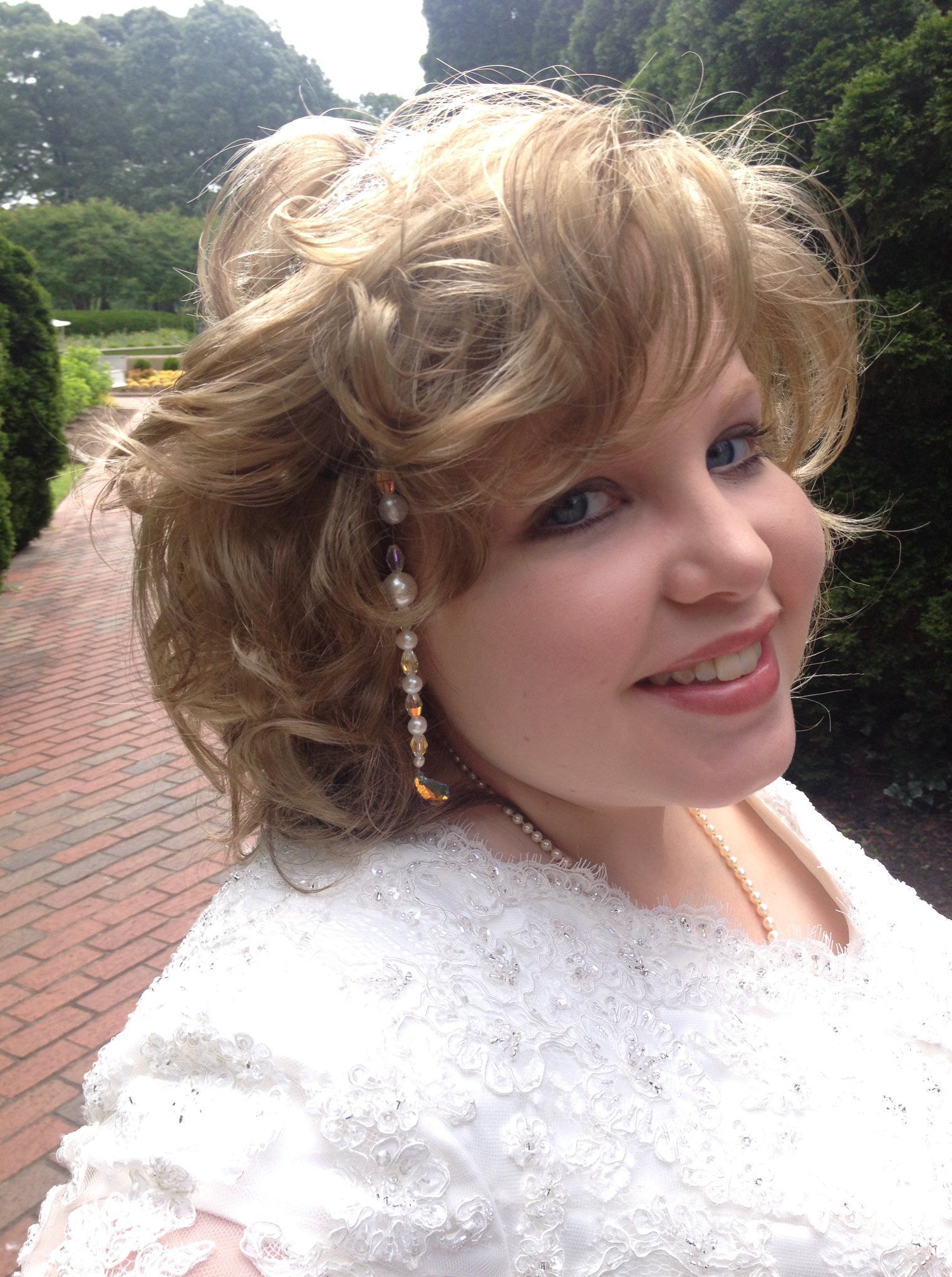 Woman with blonde curly hair, smiling, wearing a white dress outdoors, beaded earring.