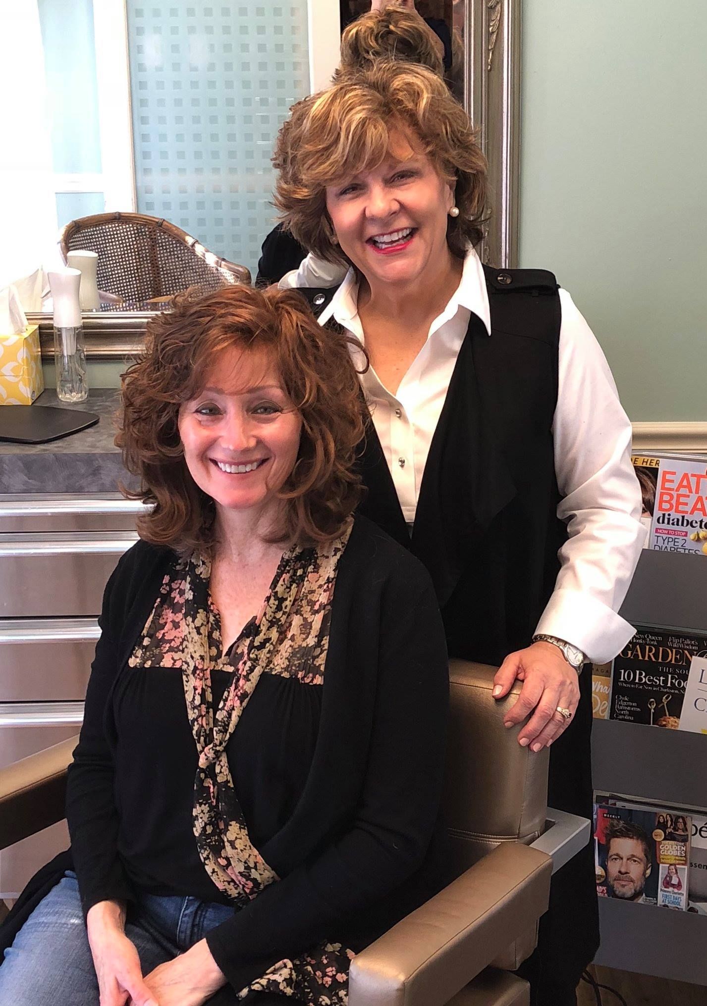 Two women smiling in a salon. One in a chair with curly hair; the other behind her, wearing a black vest.