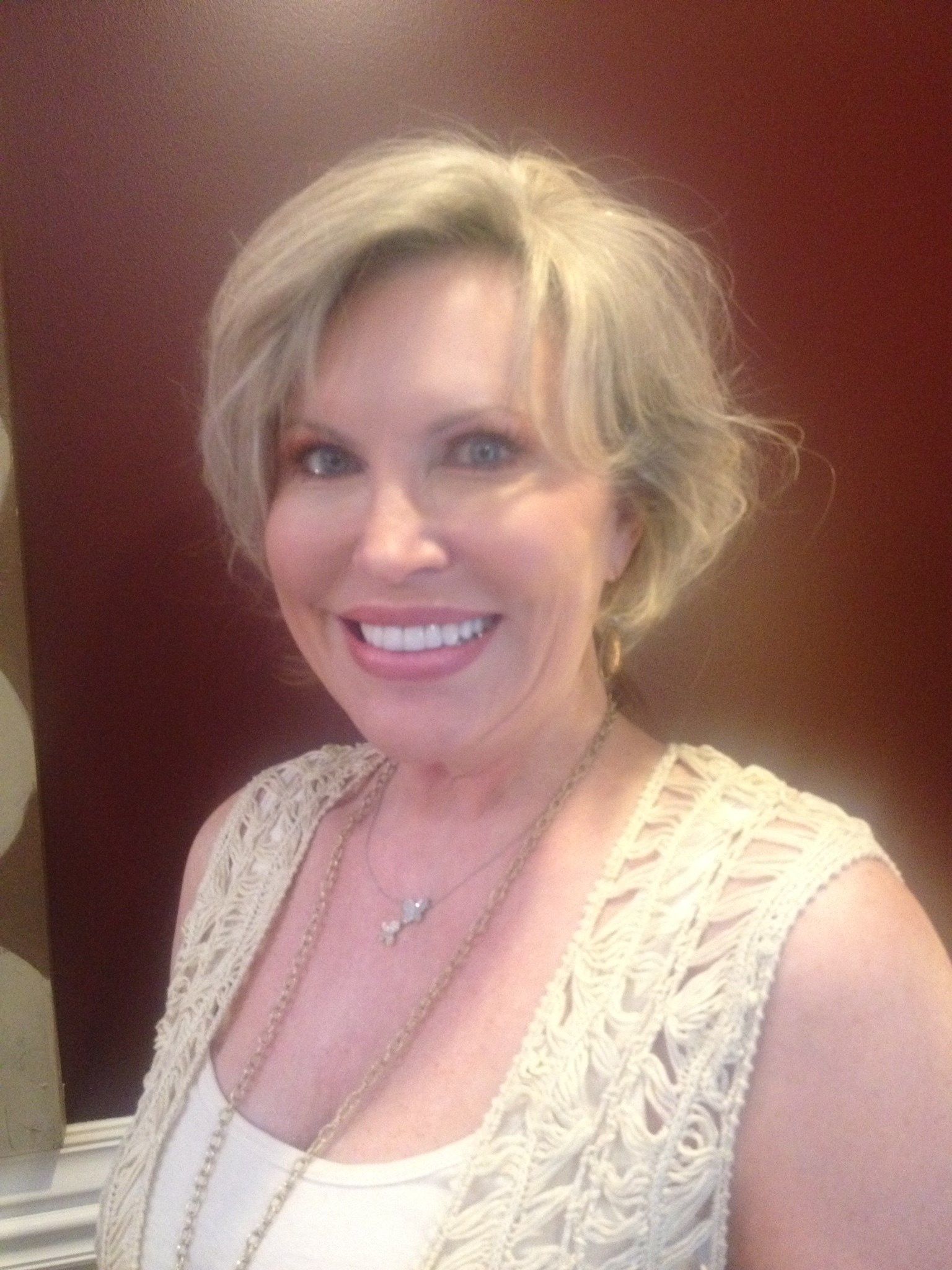 Woman with short blonde hair, smiling, wearing a cream-colored crocheted vest, necklace; red-brown background.