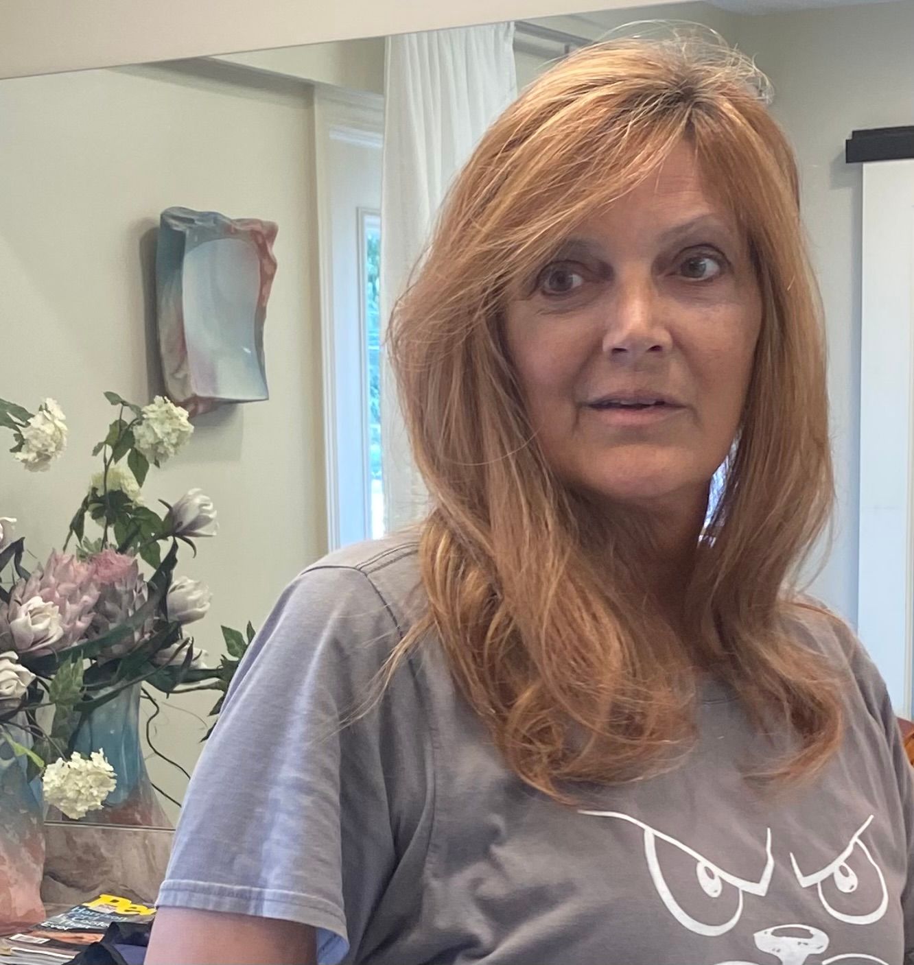 Woman with reddish-blonde hair in gray shirt looks slightly to the side, in an interior setting with flowers and artwork.