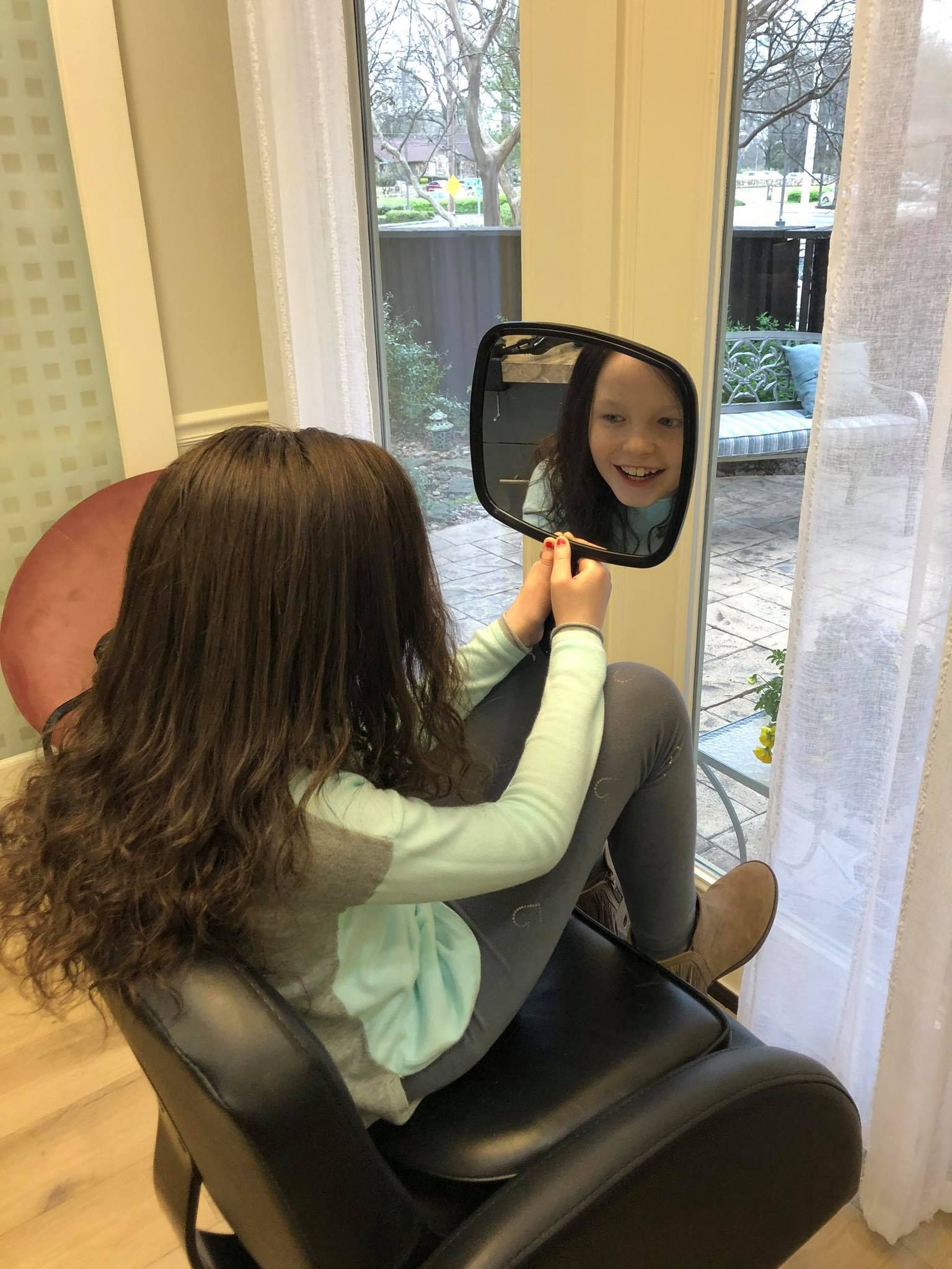 Girl in salon chair looking at herself in a hand mirror, smiling.