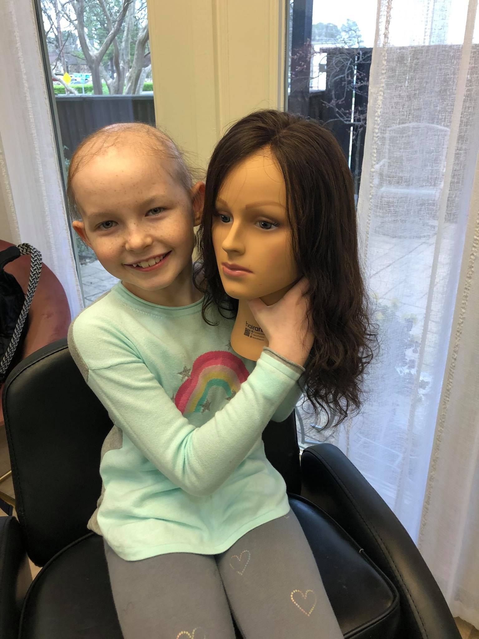 Young girl smiles, holding a mannequin head with long, dark hair. She sits in a salon chair.