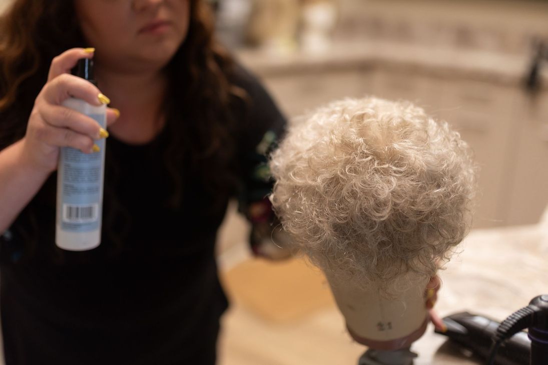 Woman spraying wig on a mannequin head, close up. Blonde, curly wig being styled in bright room.