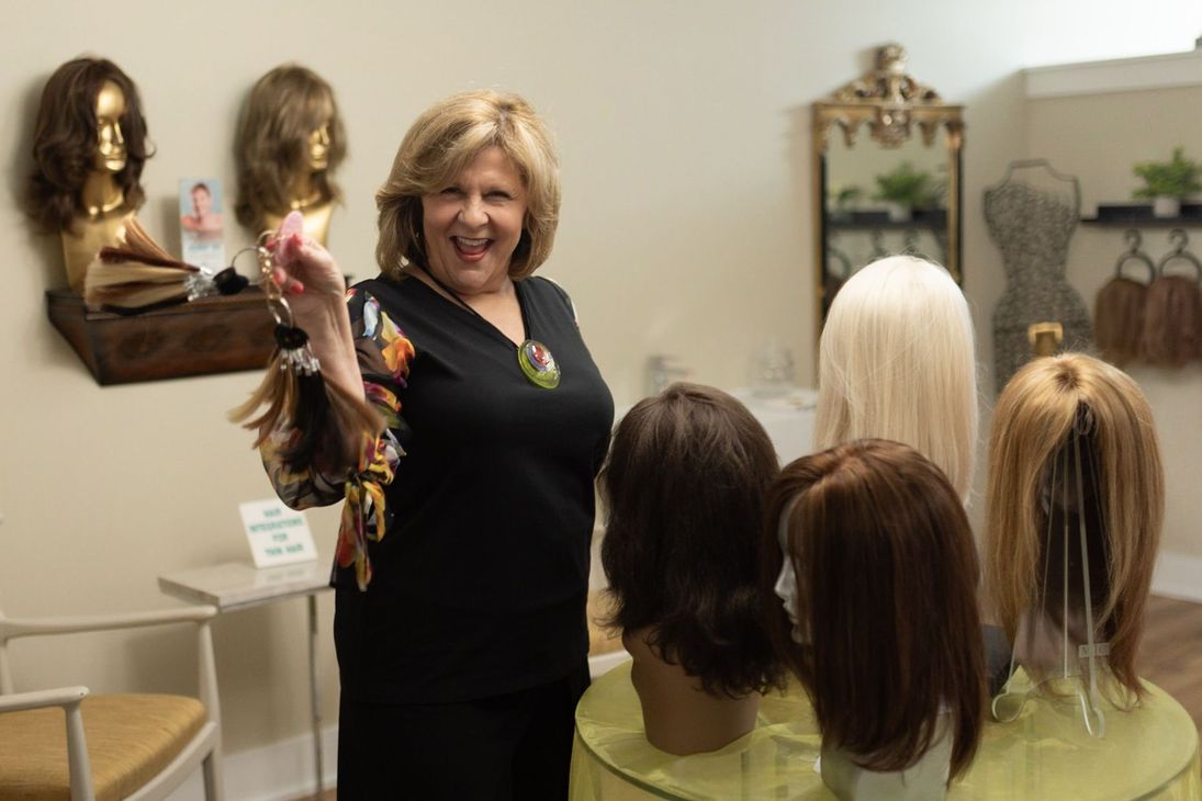 Woman in wig shop holding a wig sample, surrounded by wigs on display. Bright, warm setting.