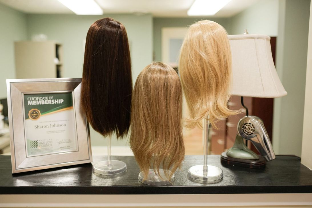 Three wigs on display: brown, light brown, and blonde, on a countertop, near a framed certificate.