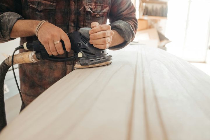 Hands sanding a light wooden plank with a power sander in a workshop.