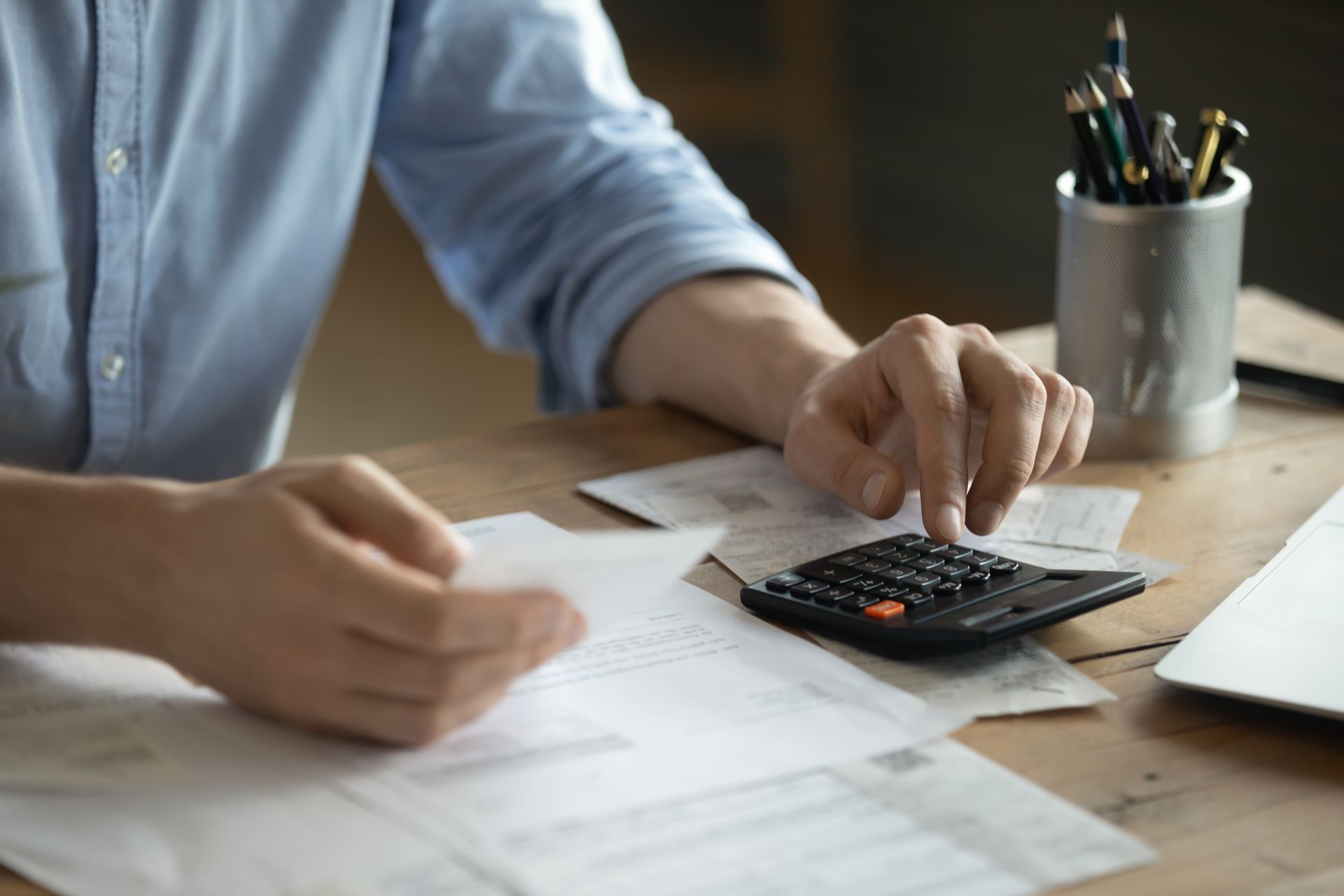 A person in a blue shirt using a calculator while reviewing financial documents at a wooden desk with a pen holder.
