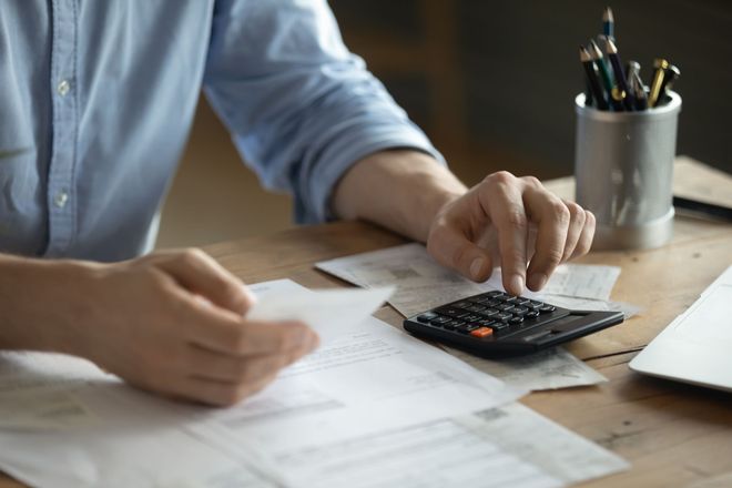 A person in a blue shirt using a calculator while reviewing financial documents at a wooden desk with a pen holder.