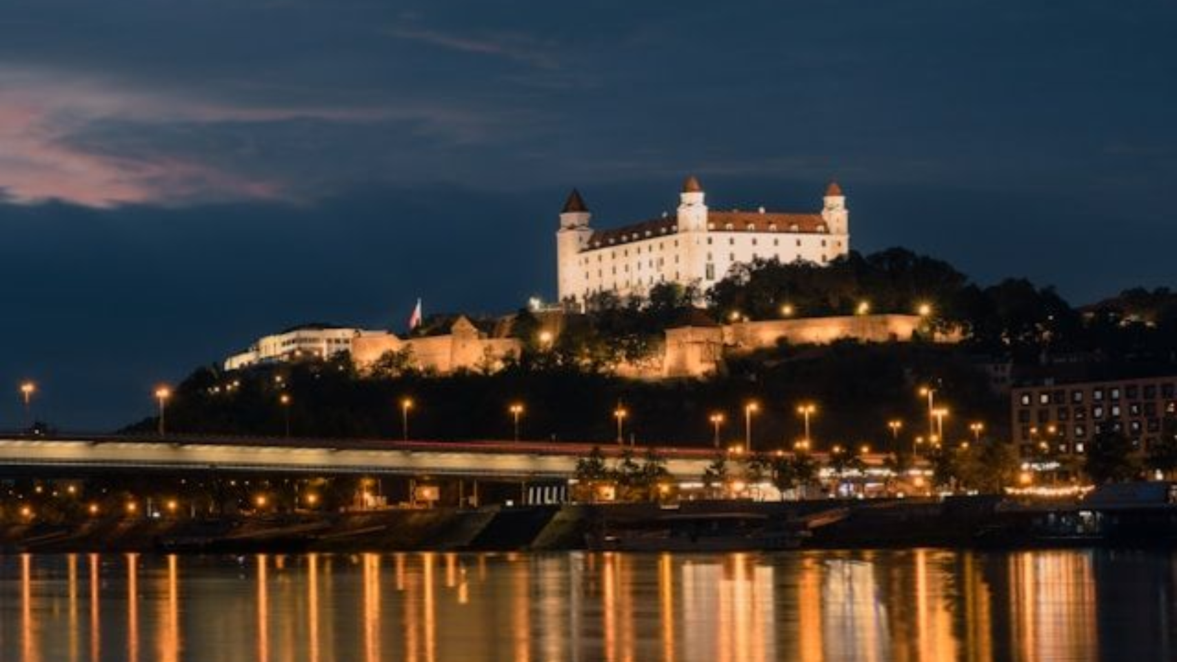 Bratislava Castle on a hill overlooking the Danube River at sunset with city lights glowing below.