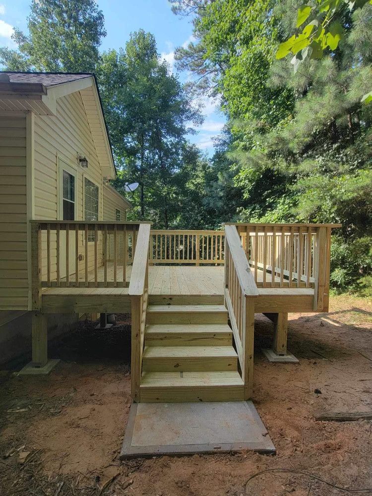 Wooden deck with stairs leading to a beige house, surrounded by trees and pine-covered ground.
