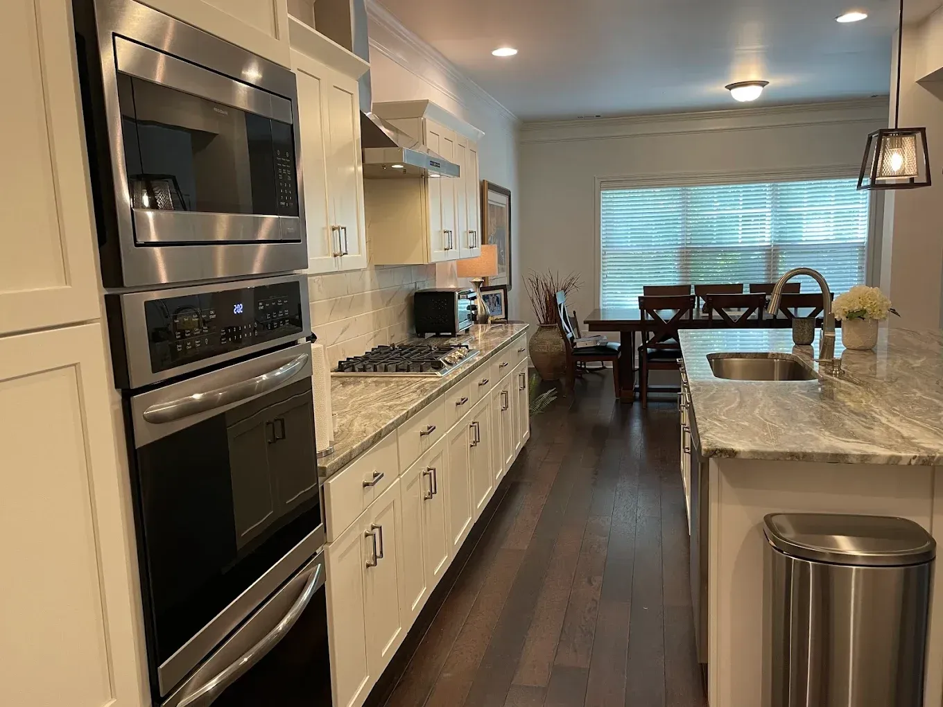 Modern kitchen with white cabinets, stainless appliances, dark wood floor, and dining area by window.