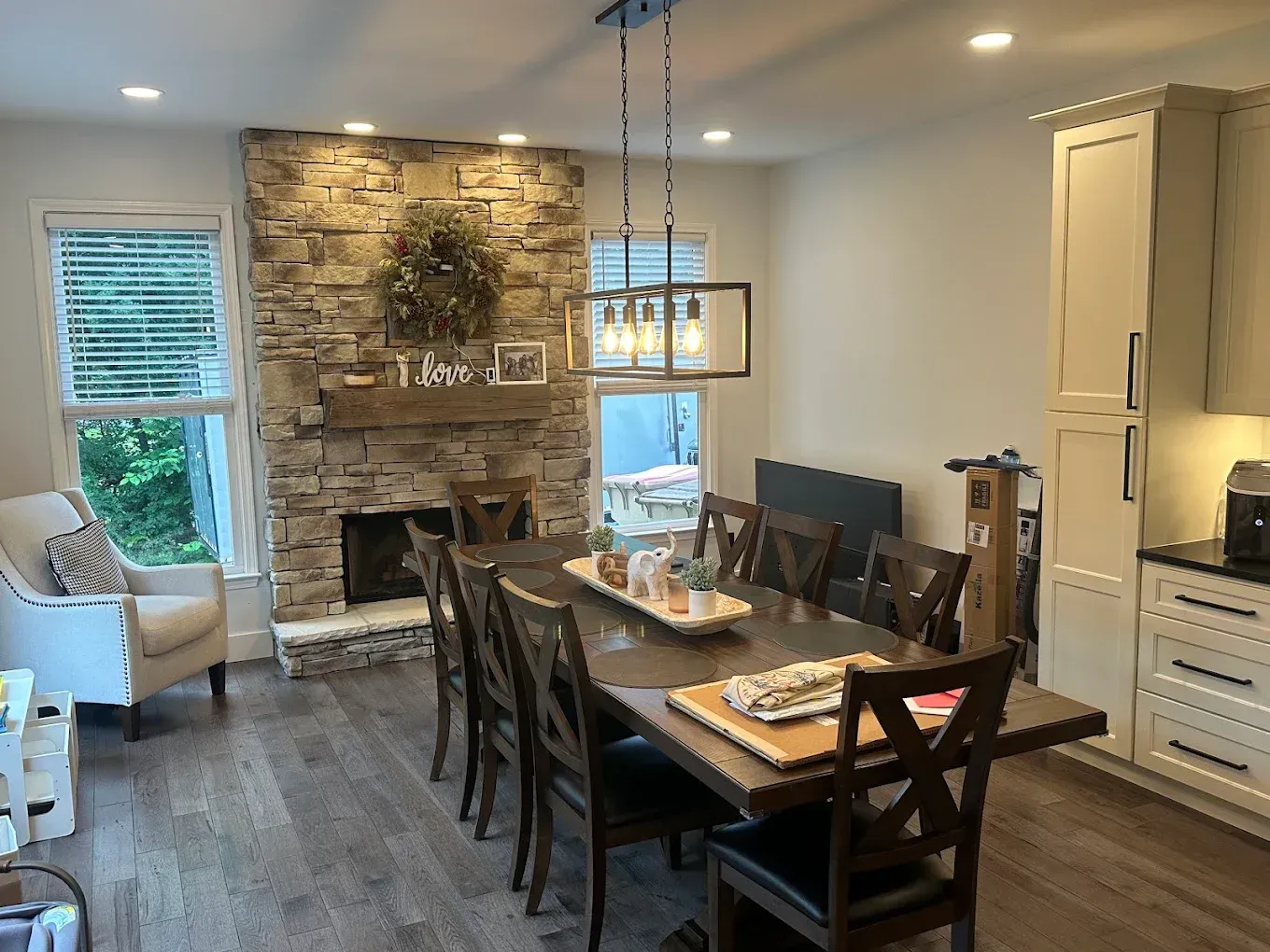 Dining room with long wooden table, chairs, stone fireplace, and bright window seating