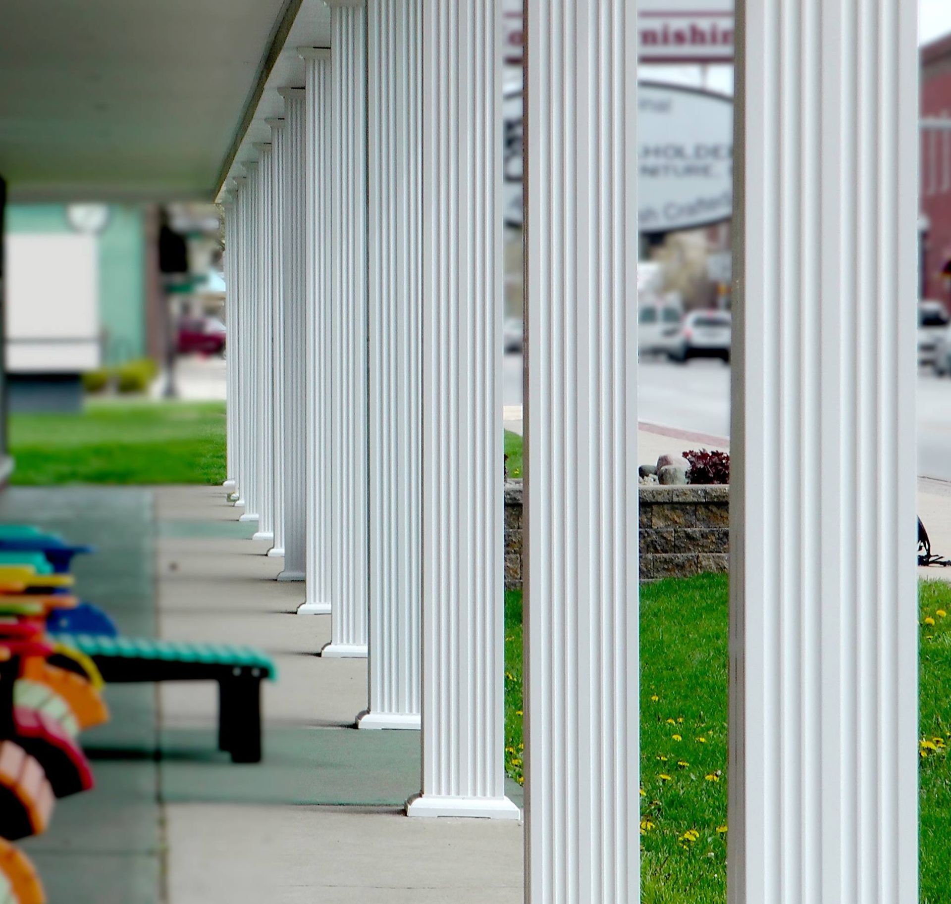 White fluted columns on a porch with colorful benches, a street, and green lawn in the background.
