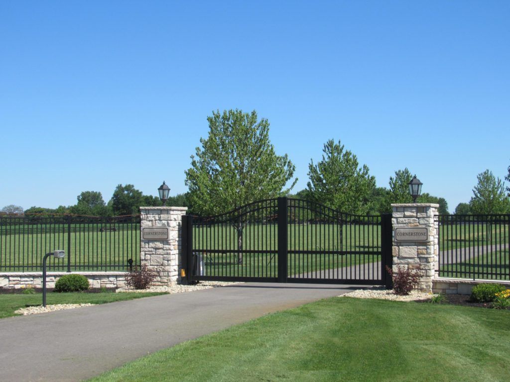 Black gated entrance with stone pillars on a paved driveway, surrounded by a metal fence and grass under a blue sky.