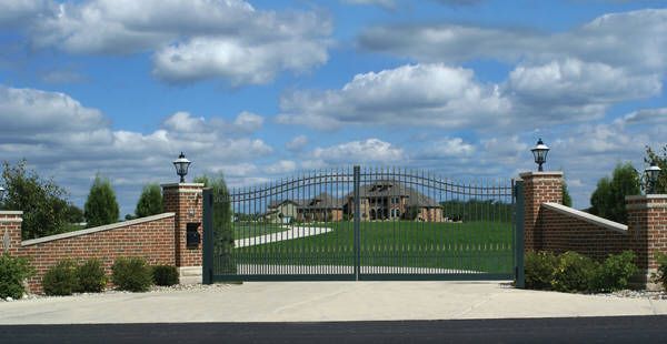 Wrought iron gates on brick pillars open to a long driveway leading to a house on a grassy lawn; blue sky with clouds.