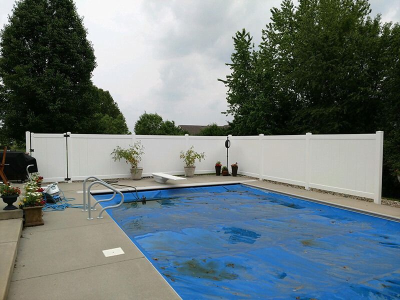 A white privacy fence encloses a backyard pool covered in a blue tarp, under a cloudy sky.