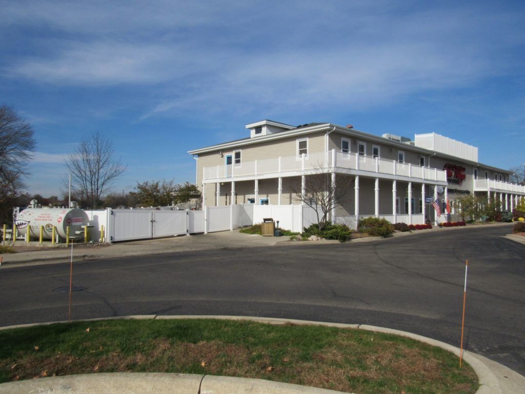 Two-story tan building with white railing, fence, and parking lot on a sunny day.