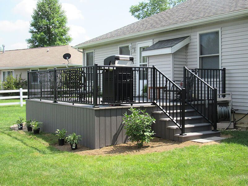 Black railing surrounds a grey deck with stairs, a house, and greenery.
