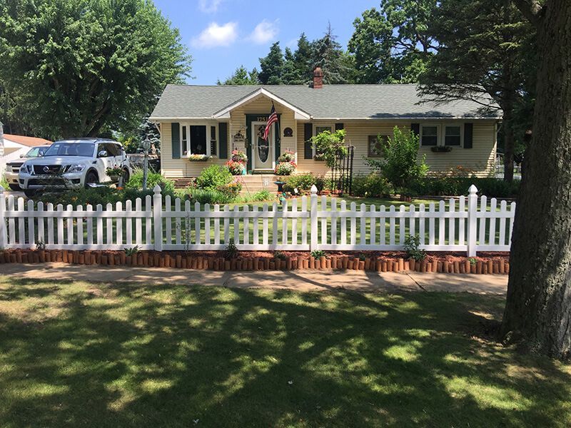 Yellow house with white picket fence, flower boxes, and a brick base in a sunny setting.