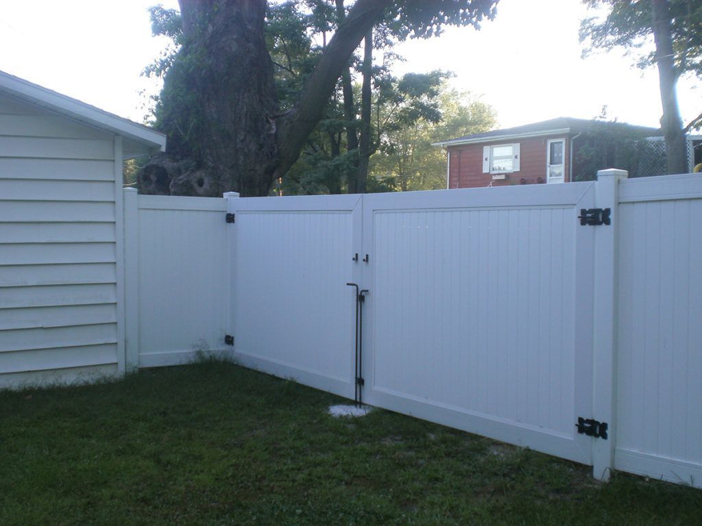White vinyl fence with gate, enclosing grassy yard. Building and trees visible in background.