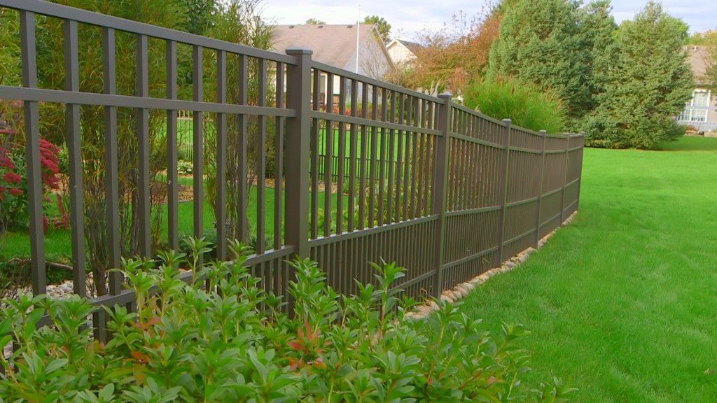 Brown metal fence curving along a grassy yard with green bushes. Houses in the background.