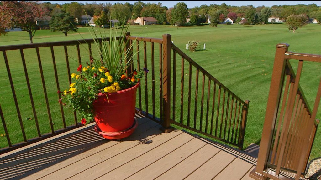 A red flower pot with yellow and orange flowers on a brown deck overlooking a green yard and houses.