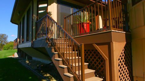 Brown deck with stairs leading to a house, lattice work, a red pot, and green lawn.