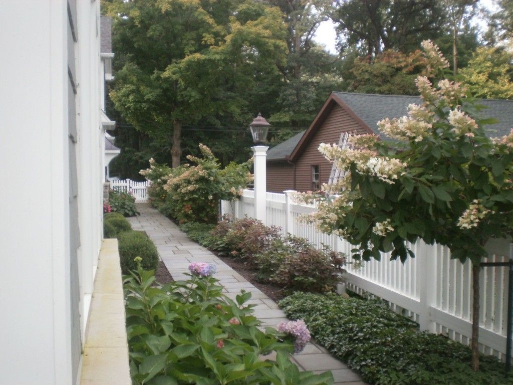 Stone pathway lined with white picket fence, bushes, and a lamp post. Building on left. Trees and a shed in the background.