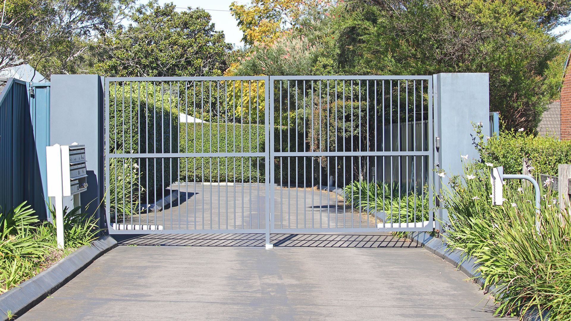 Gray metal gate spanning a driveway, flanked by pillars. Green bushes and trees in the background.
