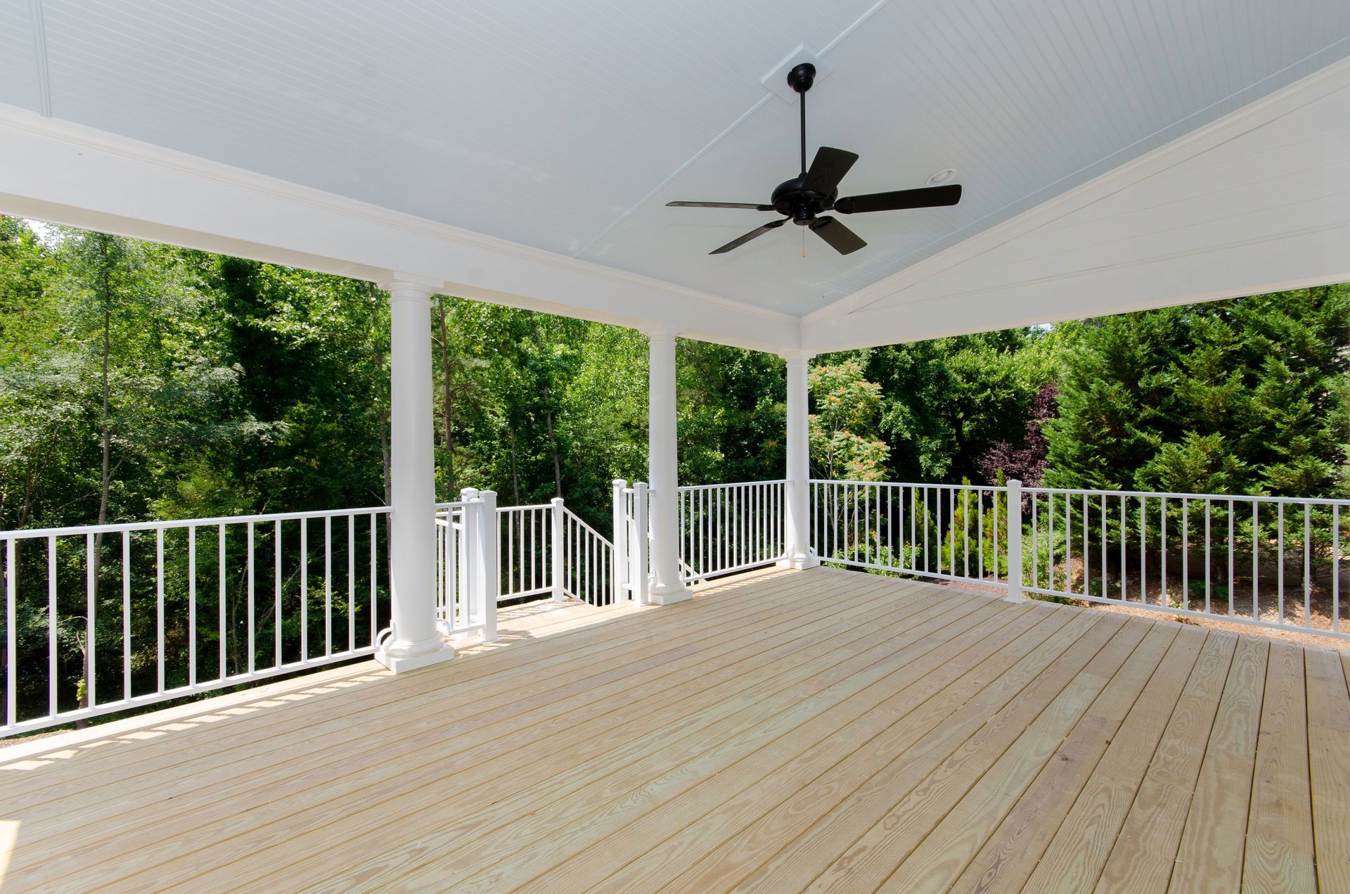 Covered wooden deck with white railing, columns, and ceiling fan; overlooking lush green trees.