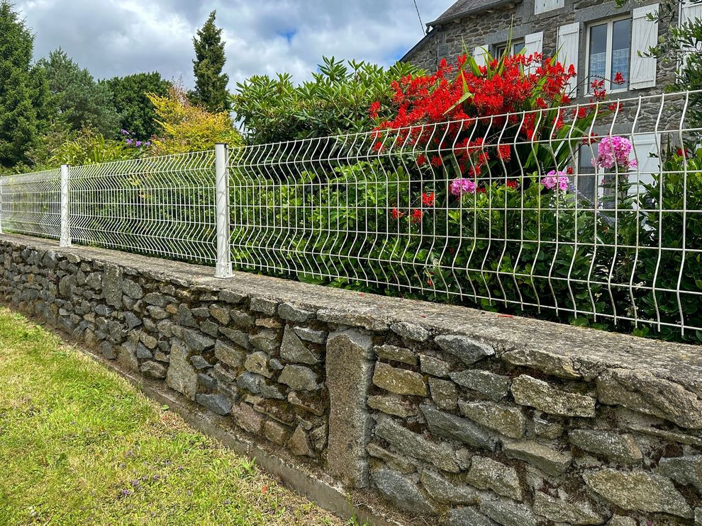 Stone wall topped with a white wire fence, greenery, and house with red and pink flowers.