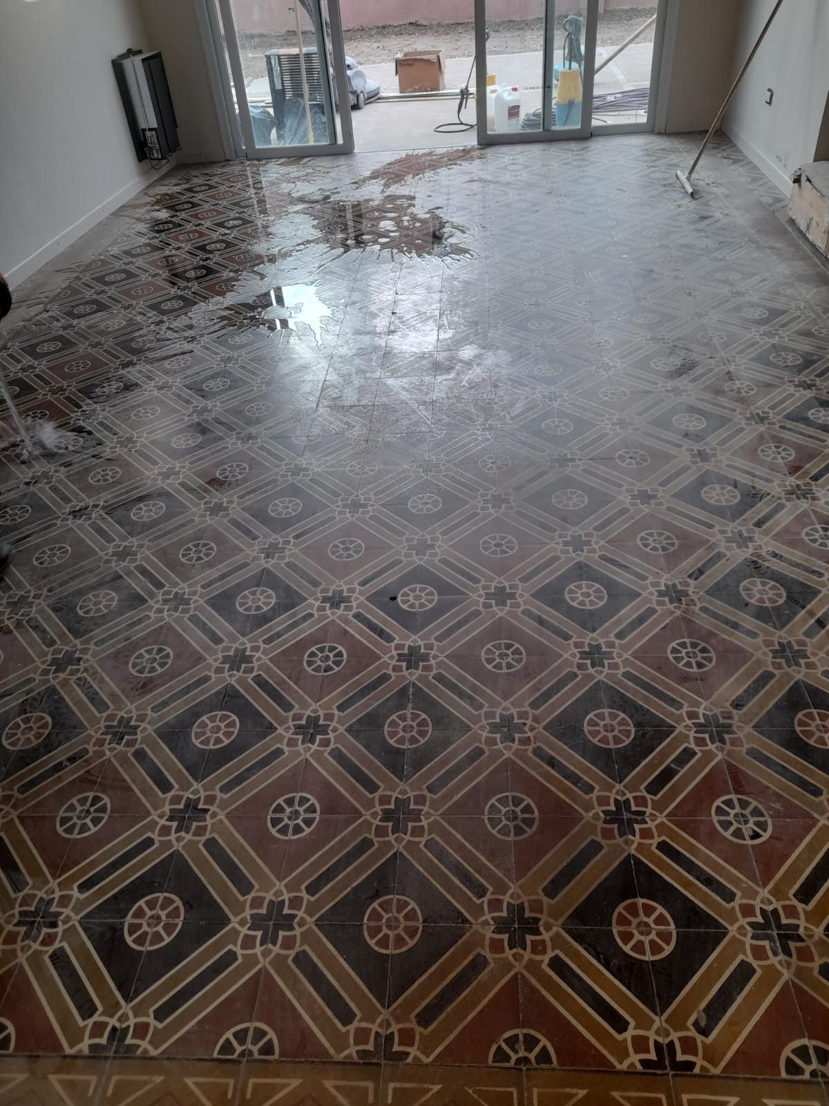 Piso mojado con diseño de baldosas oscuras, daños por agua cerca de puertas de vidrio y una pared.