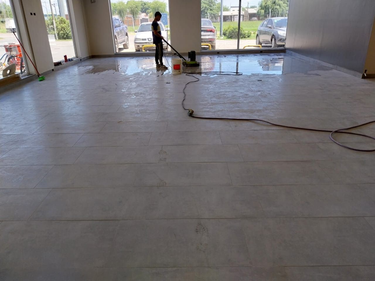 Persona limpiando un piso grande y húmedo de concreto dentro de un edificio con ventanas grandes.