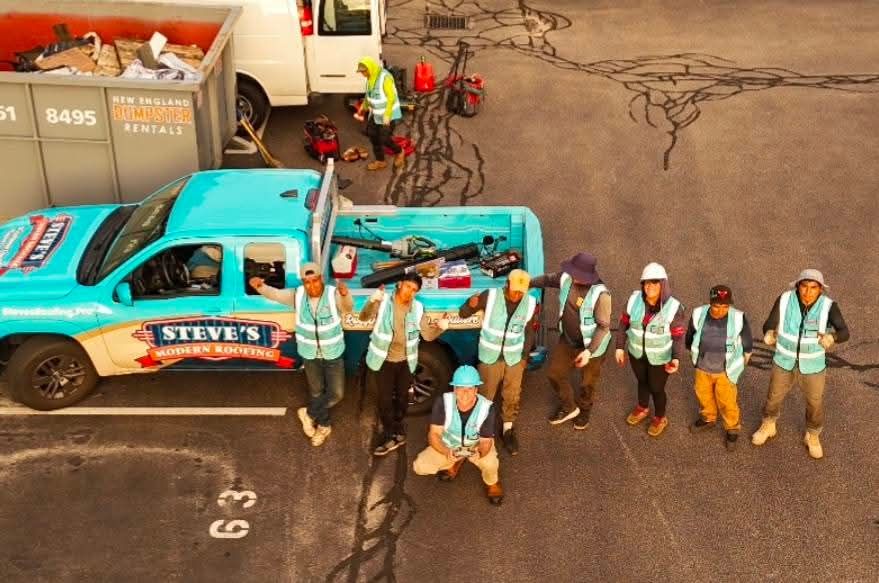 Utility workers in reflective vests stand by a turquoise truck on a wet street with cables overhead.