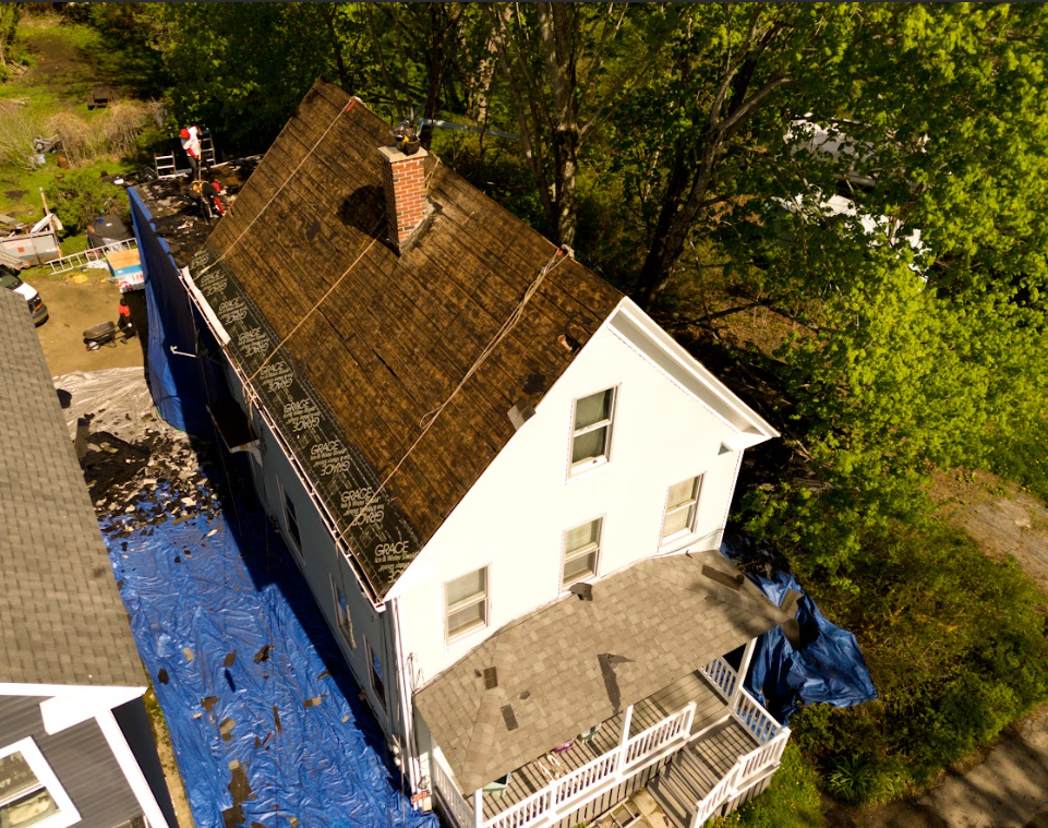 Aerial view of a white house with a brown roof beside trees, covered in blue tarp during repairs.