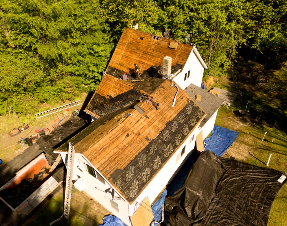 Aerial view of a house with a damaged, partially stripped roof beside dense green trees.