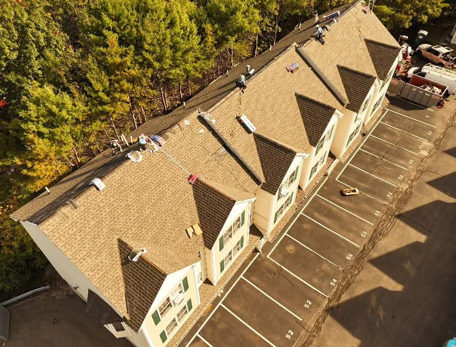 Aerial view of tan townhouses with brown roofs and parking spaces beside a wooded area