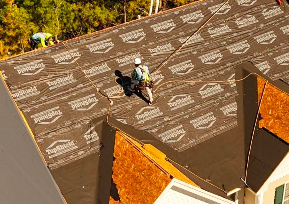 Workers installing roofing shingles on a steep roof, with roof sections visible in the foreground.