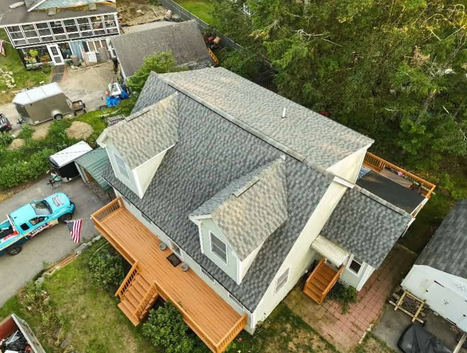 Aerial view of a gray-roofed house with a wooden deck, driveway, and parked blue car surrounded by trees.