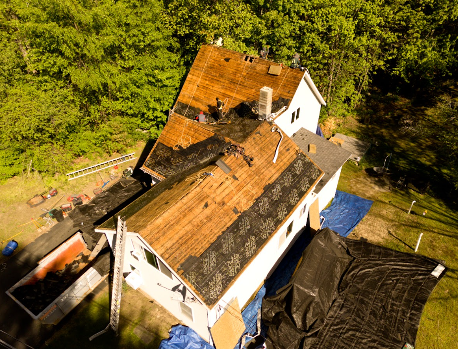Damaged shingle roof with a large exposed section and chimney under a blue sky