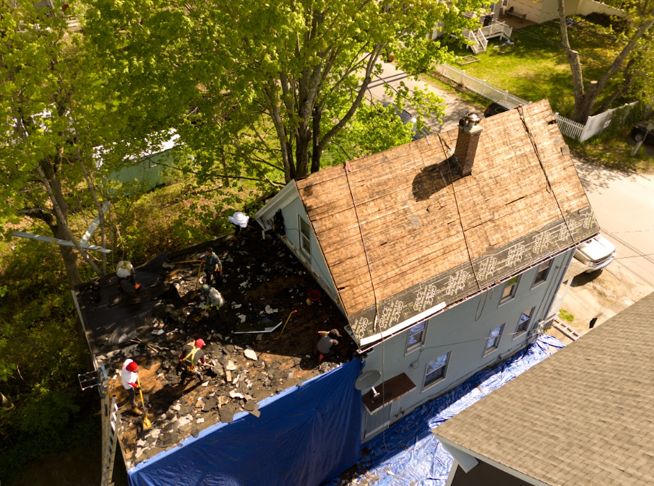 Aerial view of a house with a damaged roof beside a blue tarp-covered area and trees