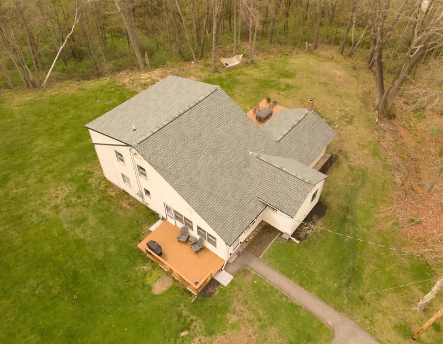 Aerial view of a beige house with gray roof, wooden deck, and walkway surrounded by a grassy yard and trees.