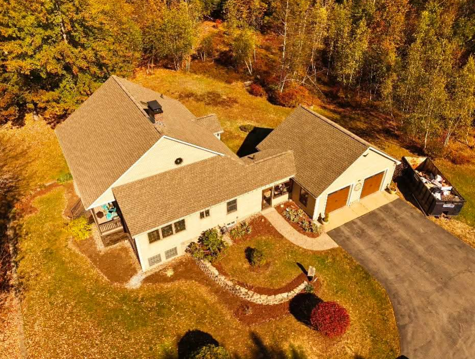 Aerial view of a beige house with a driveway, surrounded by autumn trees and a landscaped yard.