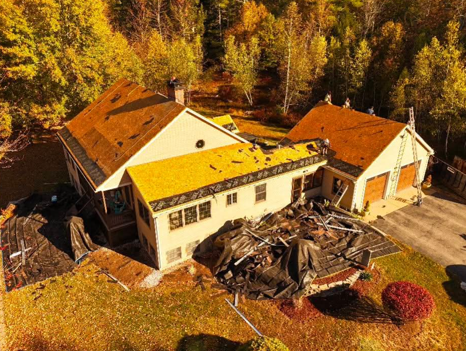 Aerial view of a house with a collapsed roof, surrounded by autumn trees and a large pile of debris.